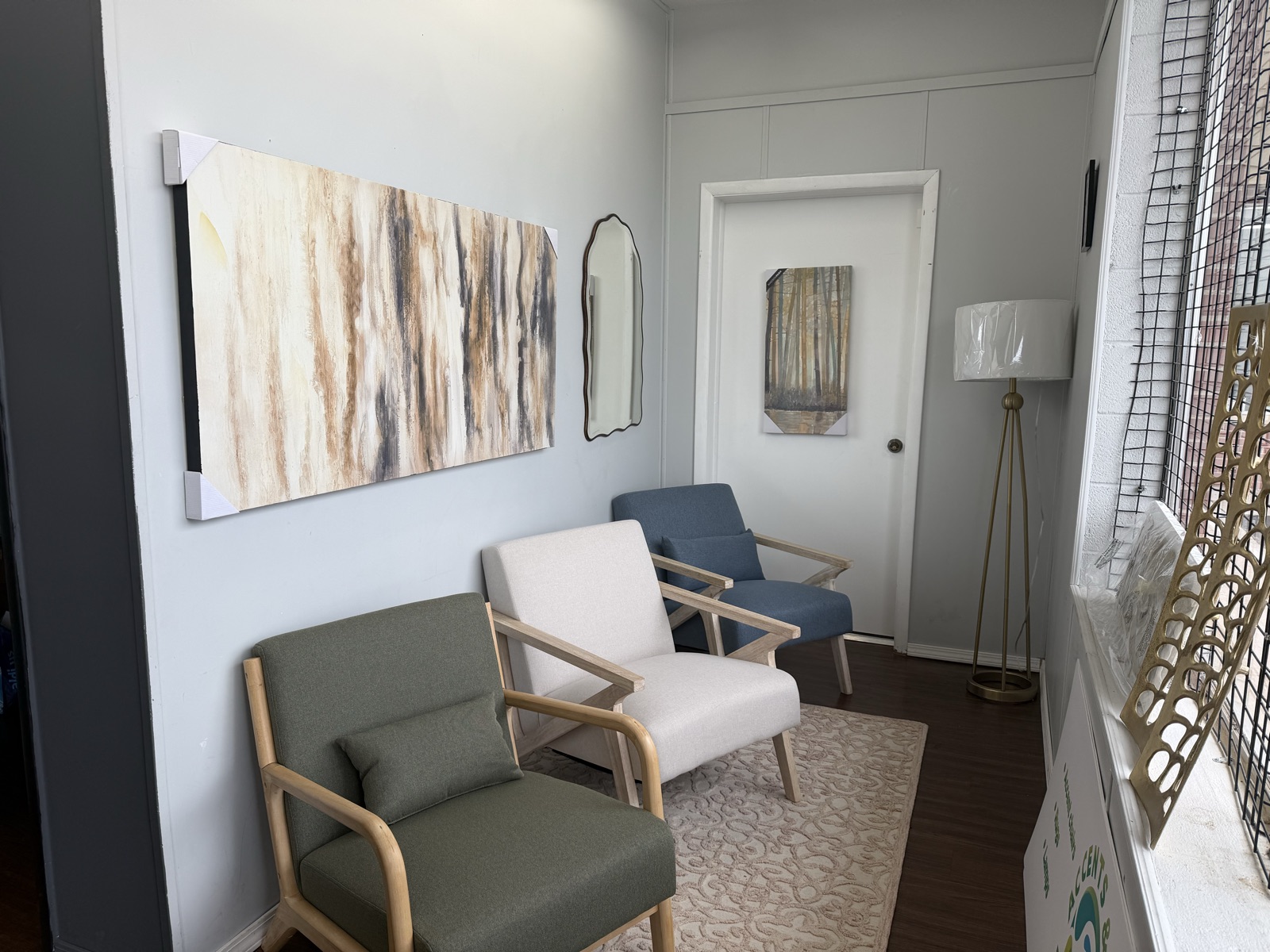 A styled corner — green and white wood-frame accent chairs and a blue armchair beneath a large abstract painting, with a mirror, floor lamp, and patterned rug.