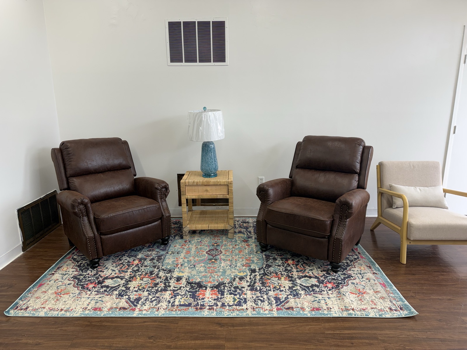 Two brown leather nailhead recliners flanking a woven side table and blue ceramic lamp, set on a blue-and-red patterned area rug.